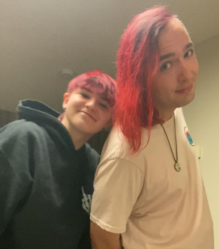 Two individuals stand close together in a softly lit indoor setting. Both have brightly colored red or pink hair, with one person wearing a black hoodie and the other in a light-colored T-shirt. The atmosphere appears friendly and casual.
