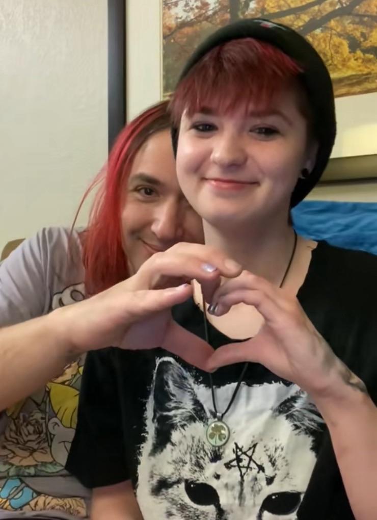 A person with red hair is peeking from behind another person, both smiling. The front person is wearing a black T-shirt with a cat design and is making a heart shape with their hands. The background features a framed picture of trees.