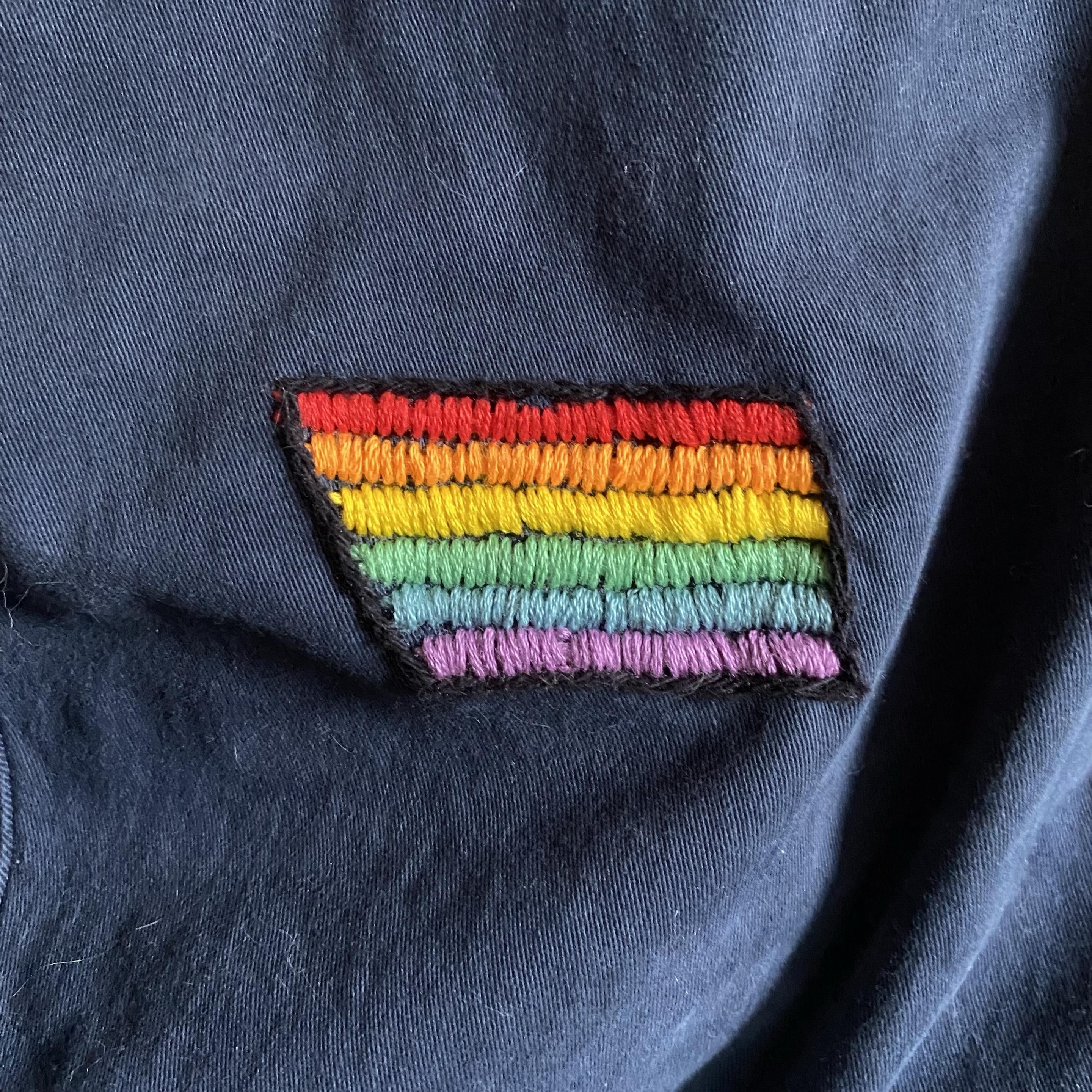 An embroidered rainbow flag patch on a dark blue jean background. The patch features horizontal stripes in red, orange, yellow, green, blue, and purple.