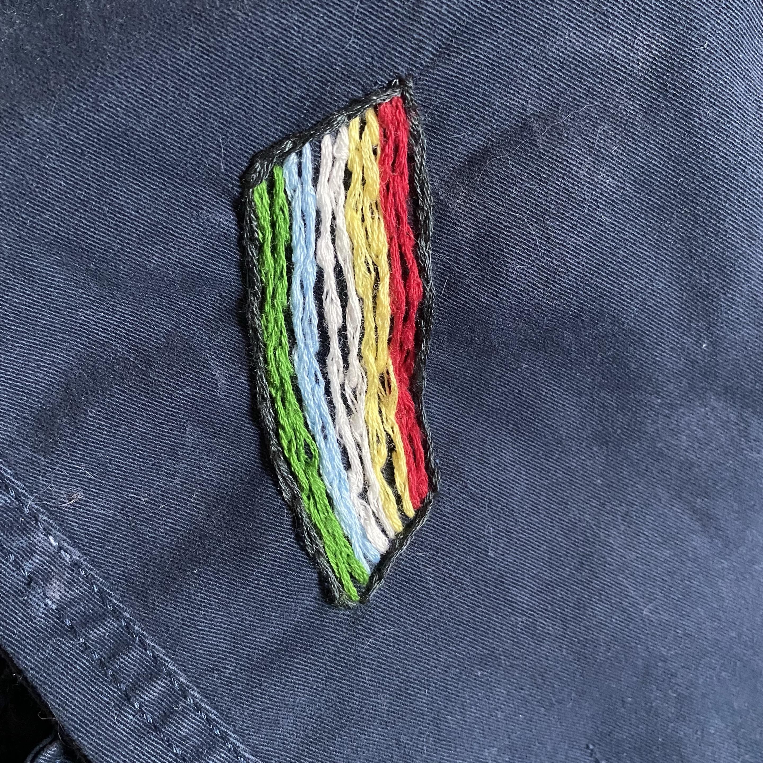 An embroidered patch featuring a stylized design with stripes of red, yellow, green, light blue, white, and black, set against a dark blue Jean fabric background. Depicting the Disability Pride flag.