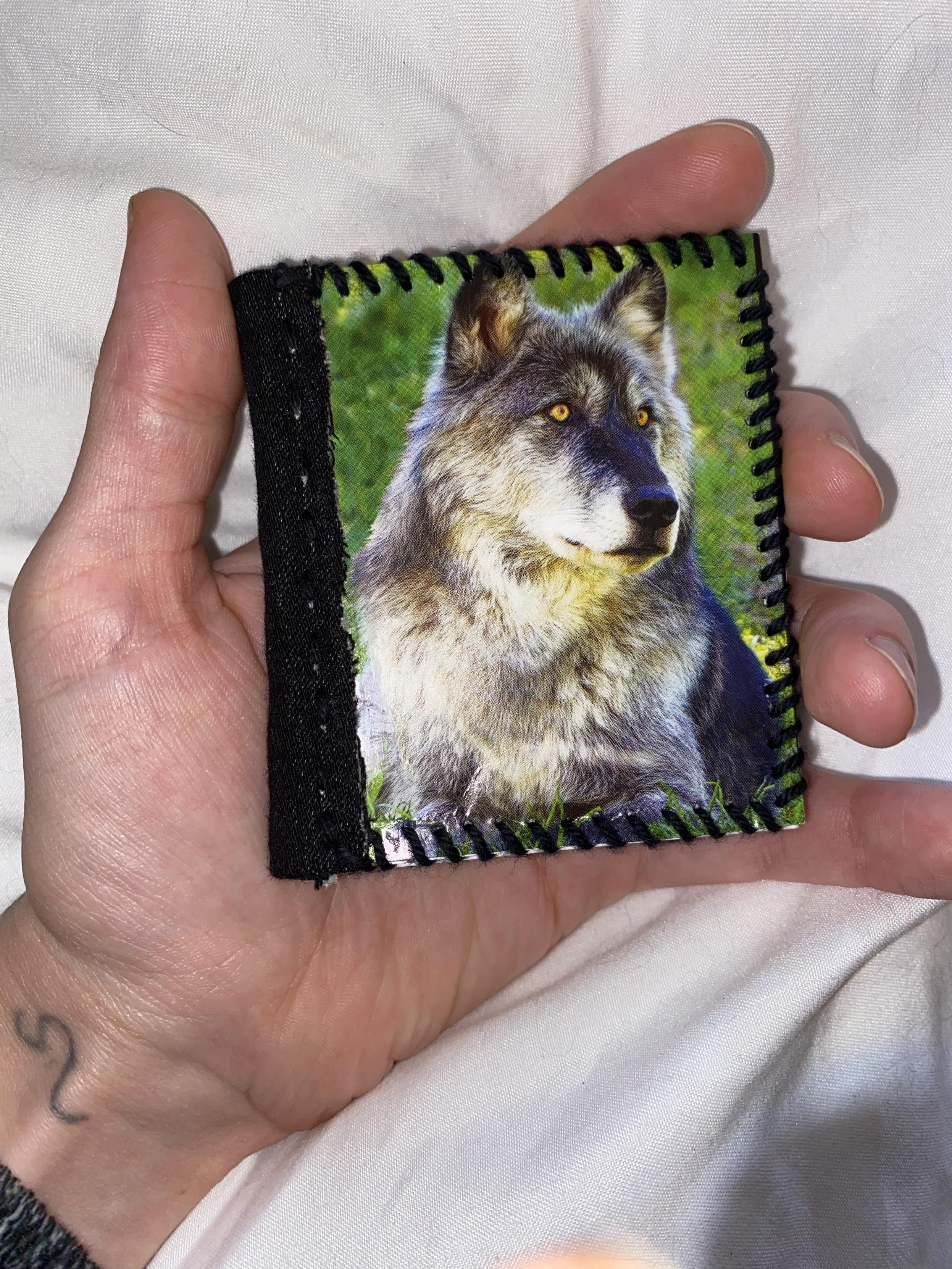 A hand holds a small, handcrafted book featuring an image of a wolf on one side, with black stitching along the edges. The background is a soft, light-colored fabric.