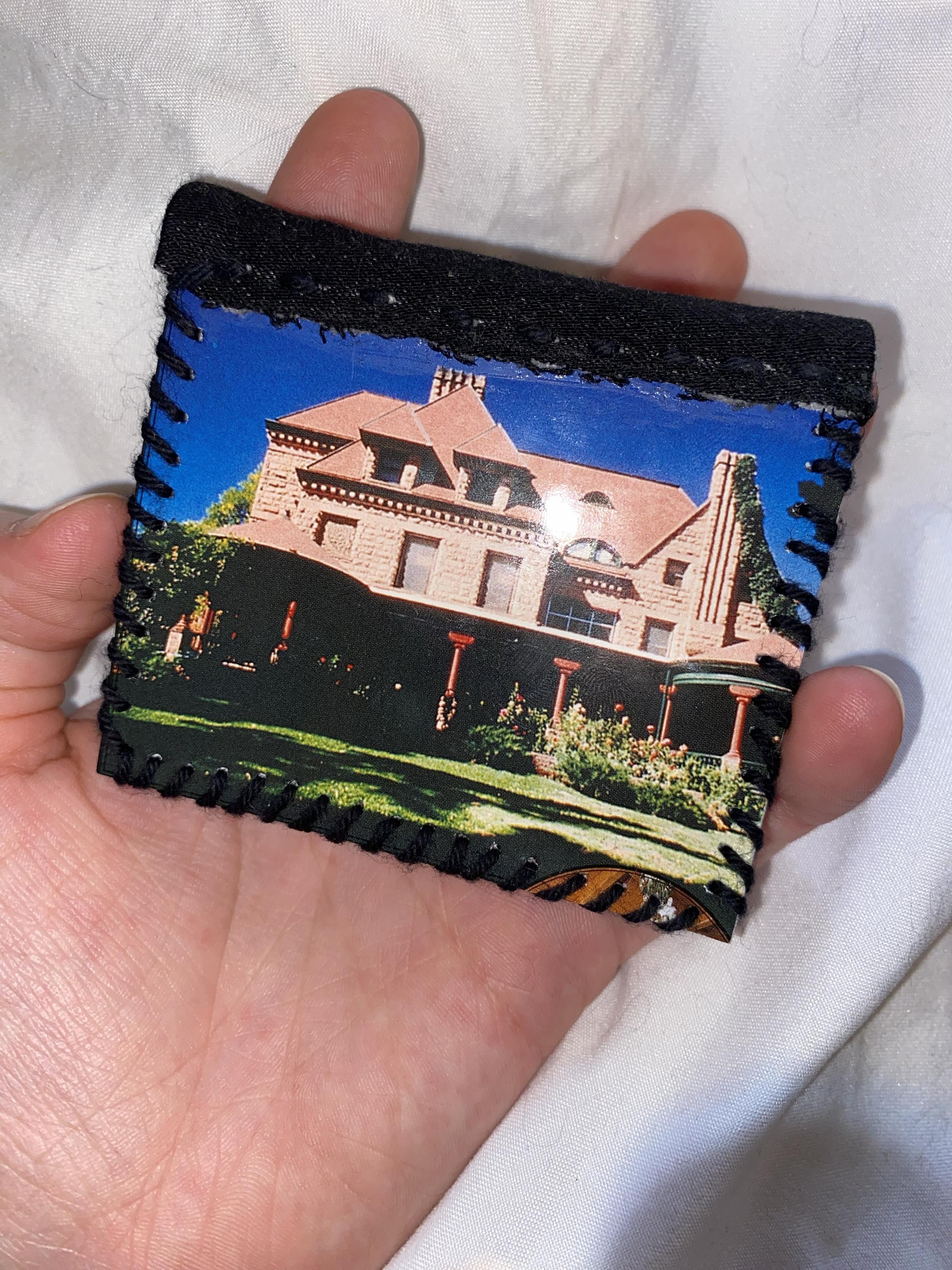 A hand holds a small, rectangular book featuring a colorful image of a large, multi-story house set in a garden. The edges are stitched with black thread, and the background is a soft, white surface.